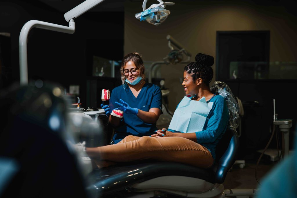 A dentist shows a dental model to a patient sitting in a dental chair; both are smiling, and dental equipment is visible in the room.