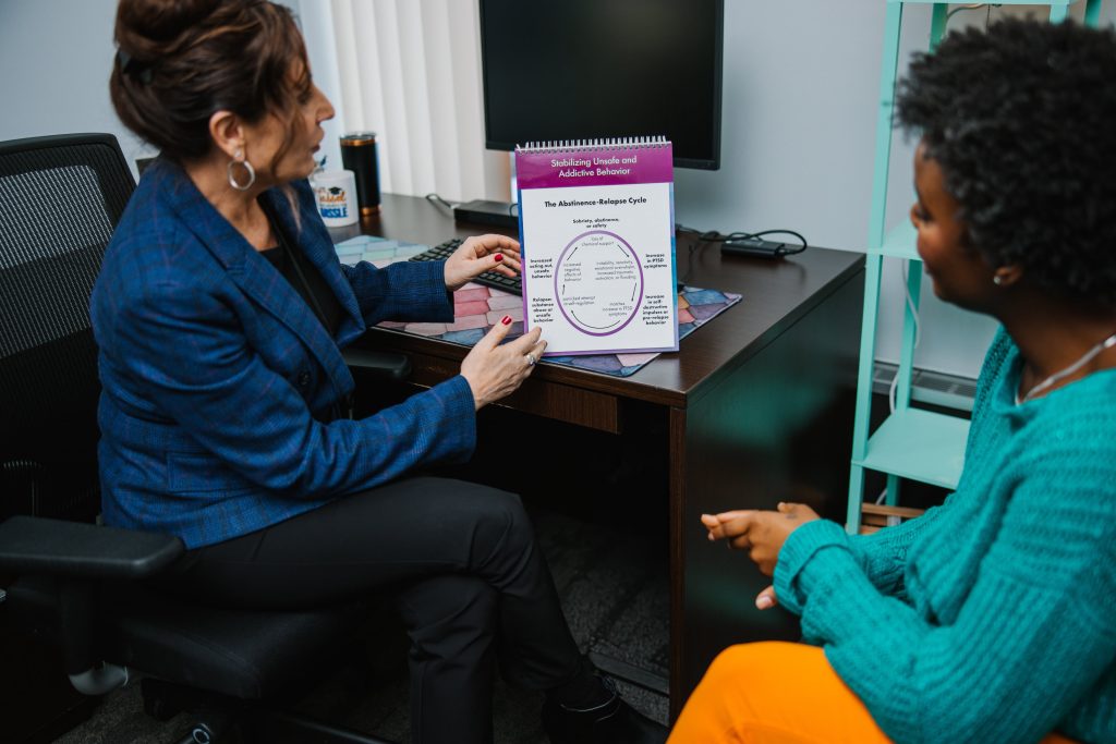 Two women sit in an office; one points to a diagram titled "The Relapse/Relapse Cycle" on a flipchart while the other listens attentively.